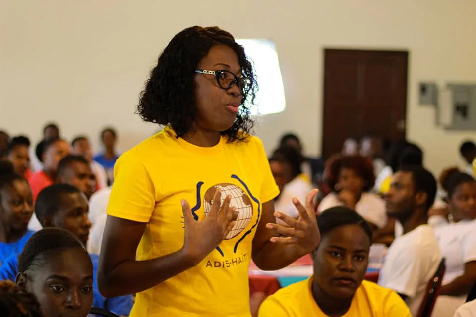 Une intervenante en t-shirt jaune ADIS-HAÏTI s'adresse à une salle remplie de jeunes participants