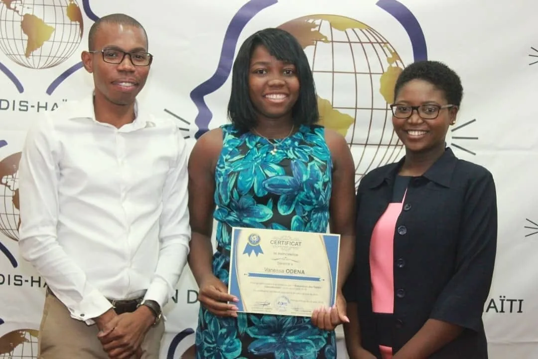 Une participante tient son certificat, entourée de deux responsables, devant un fond aux couleurs d'ADIS-HAÏTI