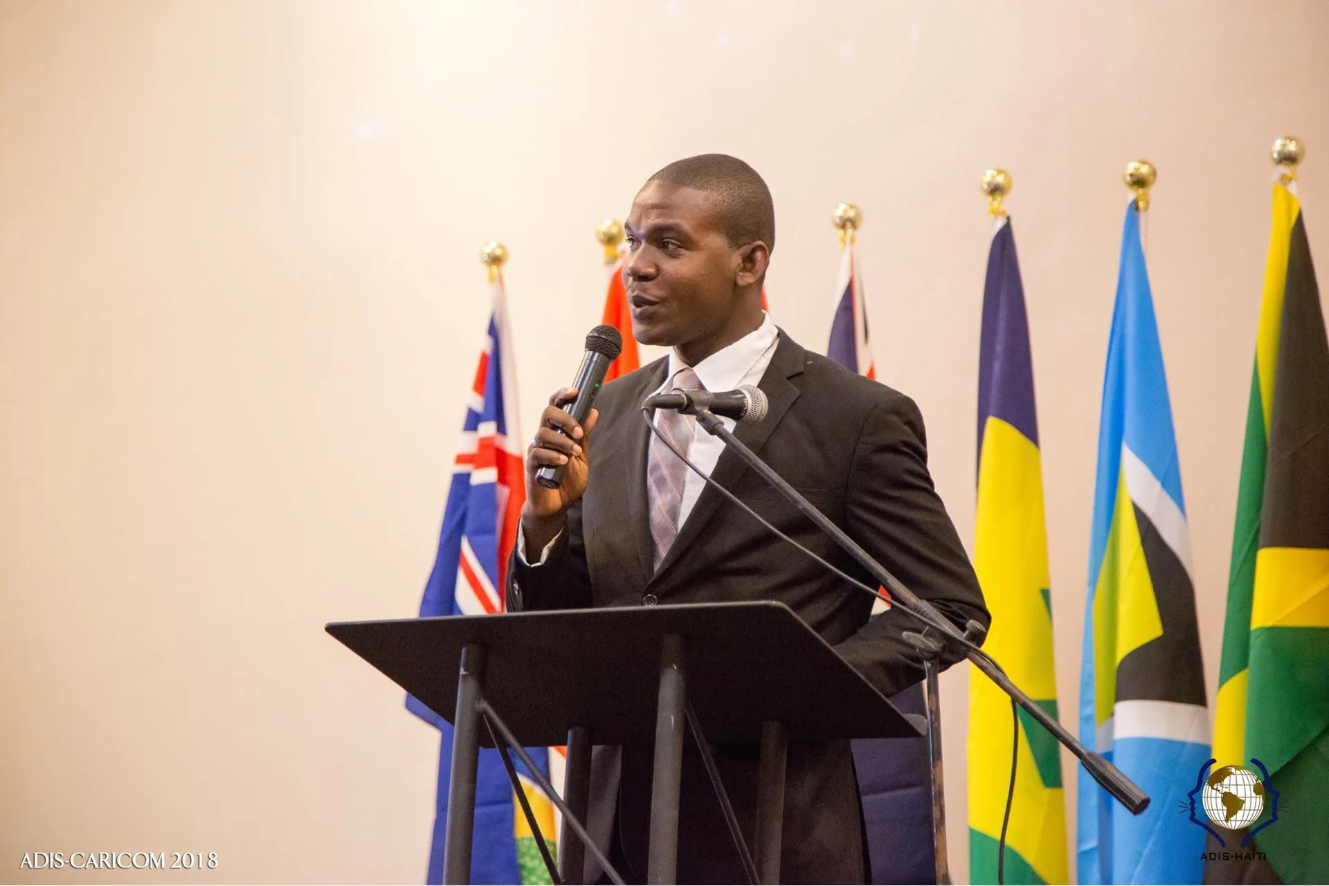 Un intervenant d'ADIS-HAÏTI s'exprime au pupitre devant les drapeaux des pays de la CARICOM, dont celui d'Haïti, lors d'ADIS CARICOM 2018