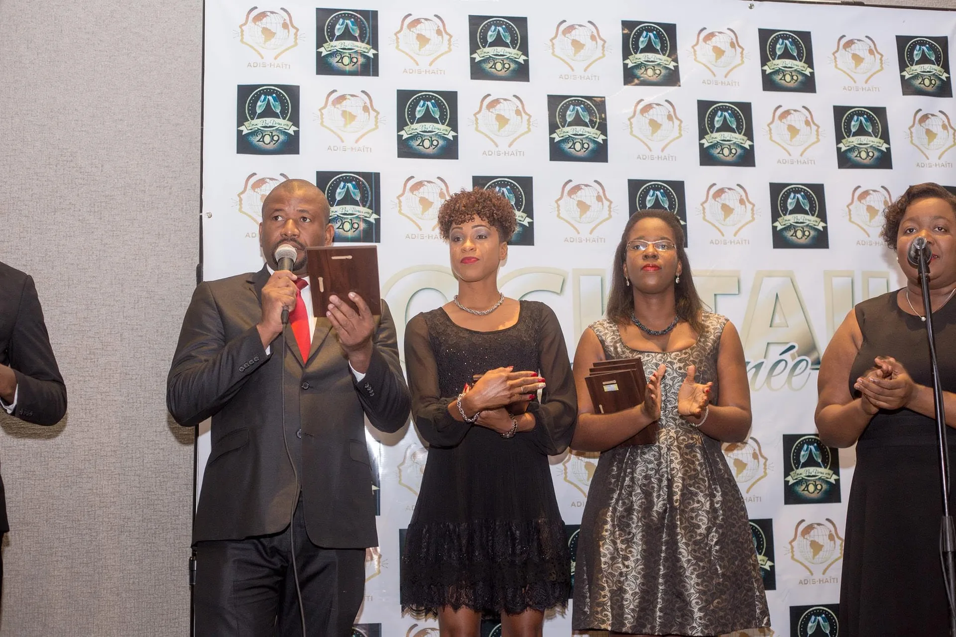 Remise de plaques d'honneur lors d'une soirée d'ADIS-HAÏTI, devant un mur aux logos de l'organisation